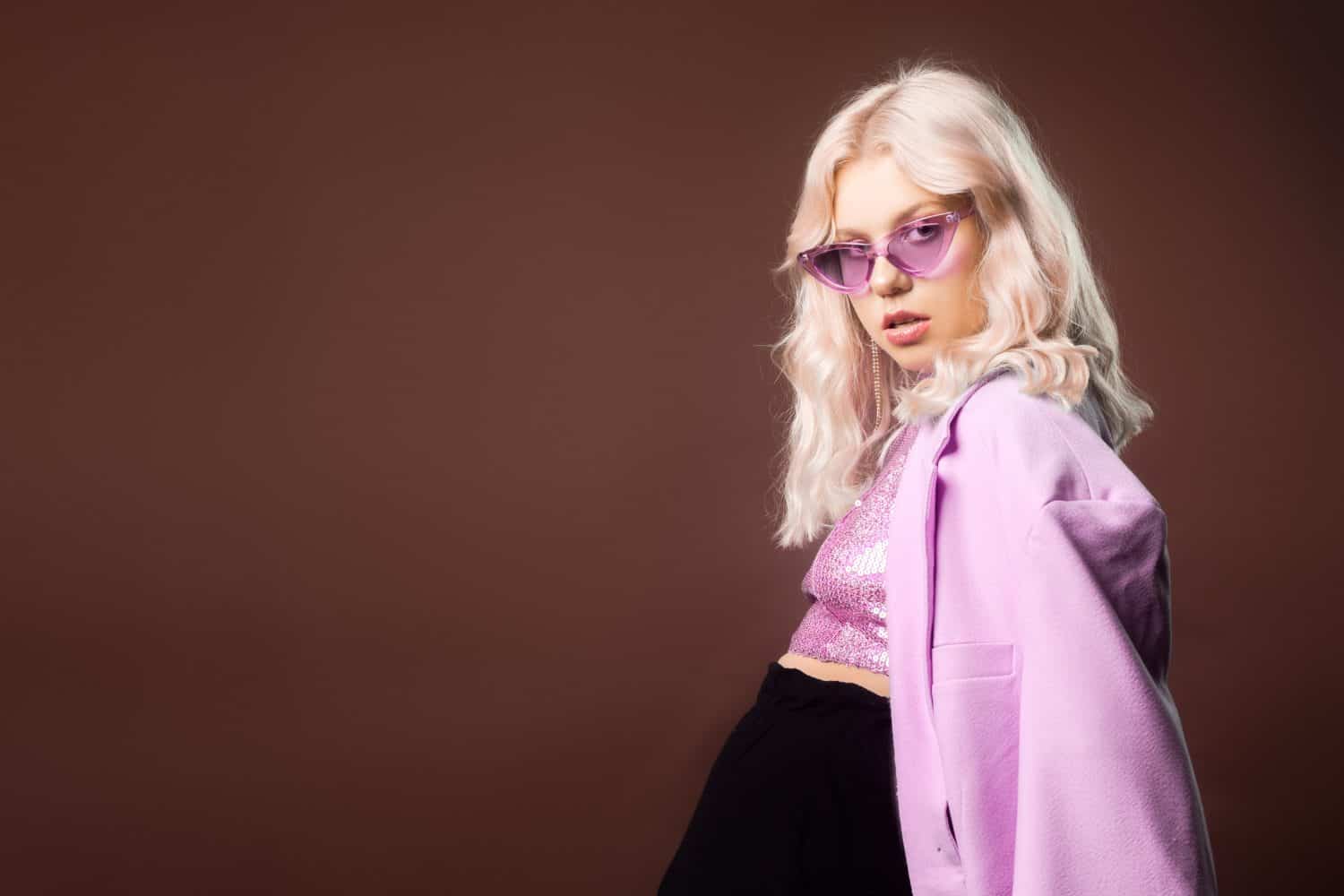 Blonde in a pink jacket with glasses, a jacket with sequins posing in the studio on a brown background. High fashion, 90s 80s. The girl dances and moves. Model