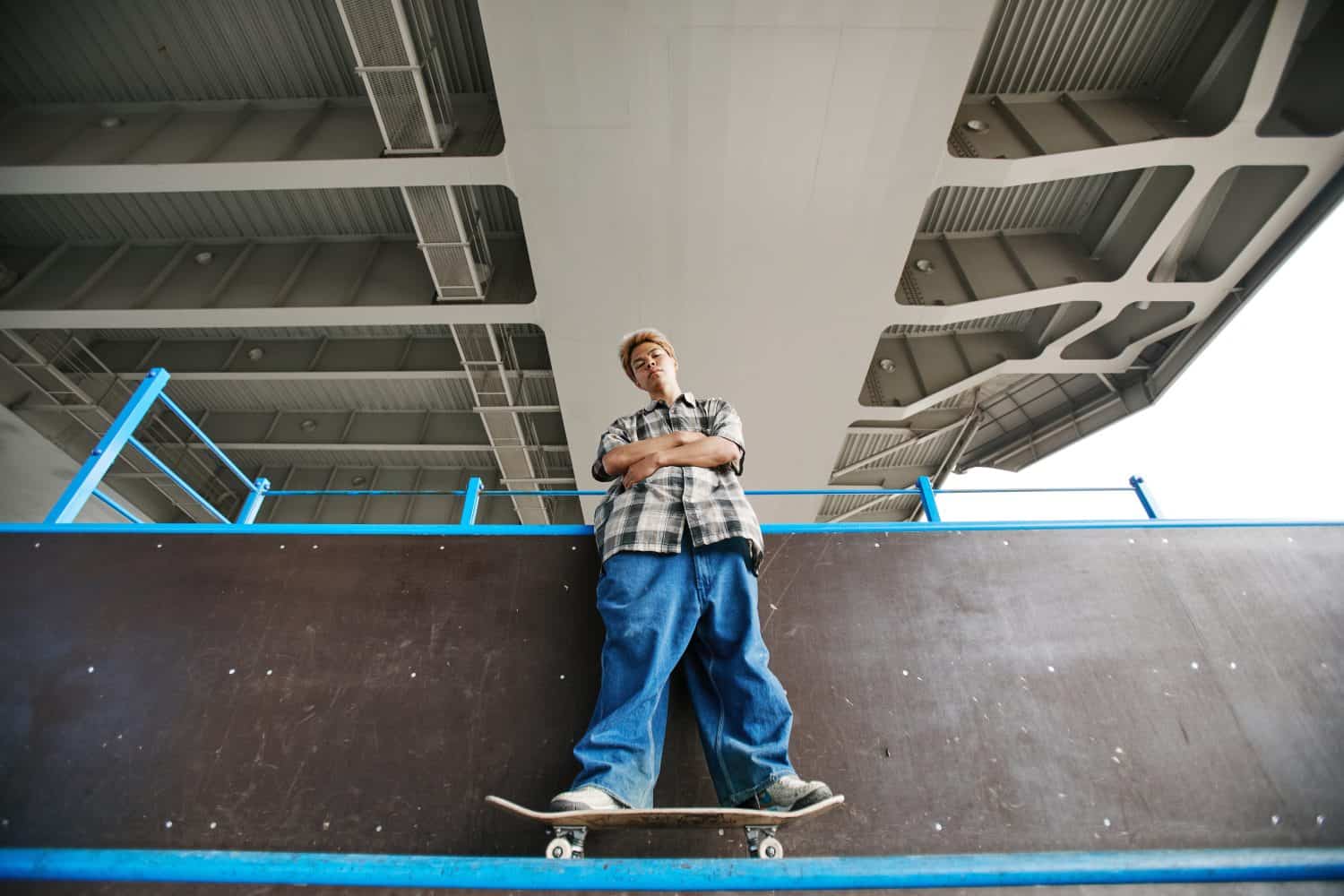 Low angle portrait of teenage boy wearing baggy pants standing on skateboard in urban area and looking at camera, copy space