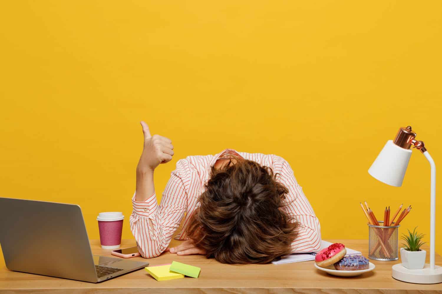 Young sleepy employee business woman wear casual shirt sit work at office with pc laptop put head down on desk sleep show thumb up isolated on plain yellow color background Achievement career concept