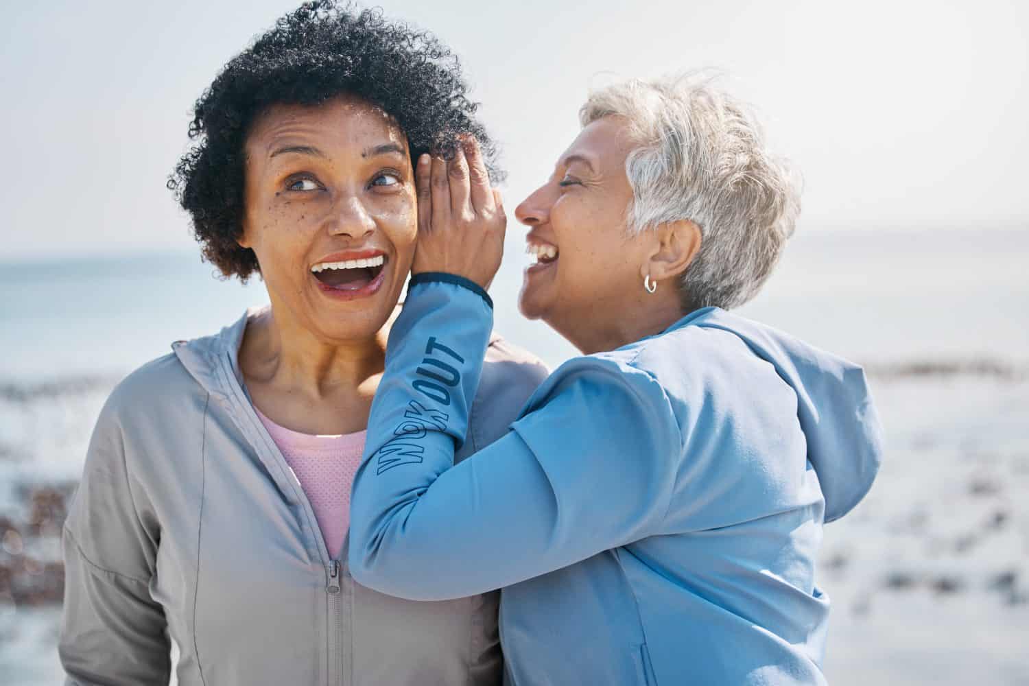 Gossip, beach and senior friends with a secret, whisper or talking in ear for a funny joke after outdoor exercise. Laughing, crazy and elderly women listening to conversation or story at the sea
