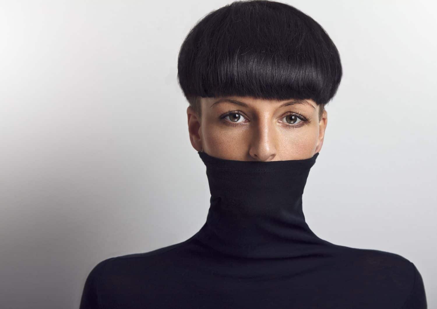 Beautiful woman with black mushroom hair cut covering her face with black turtleneck looking at the camera.