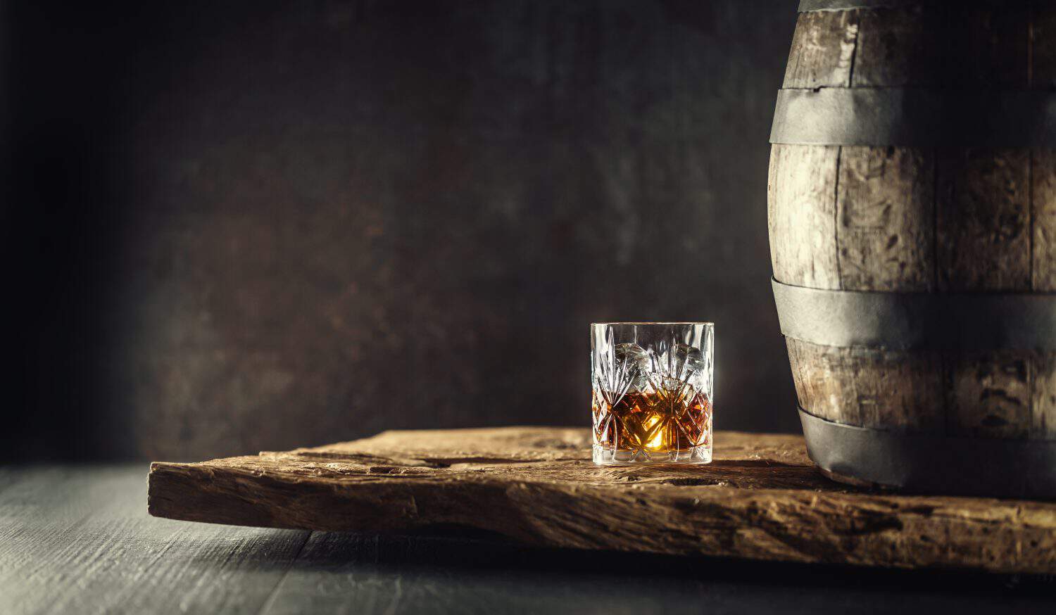 Glass of whisky cognac or bourbon in ornamental glass next to a vinatge wooden barrel on a rustic wood and dark background.