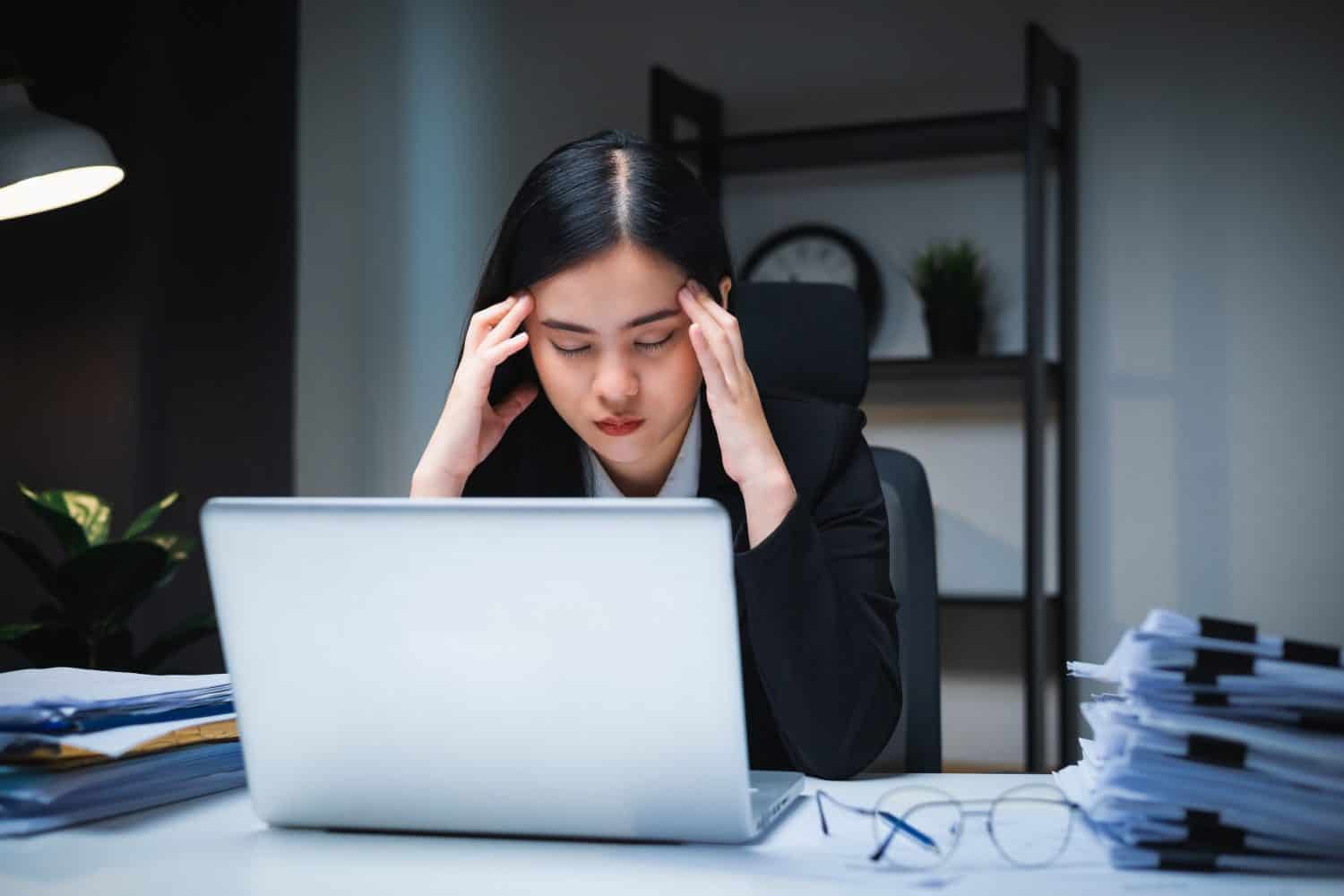 Stressed business woman working late at night in the office hands on head feeling headache. Tired woman looking at laptop working hard sitting in the dark room office. Overtime concept