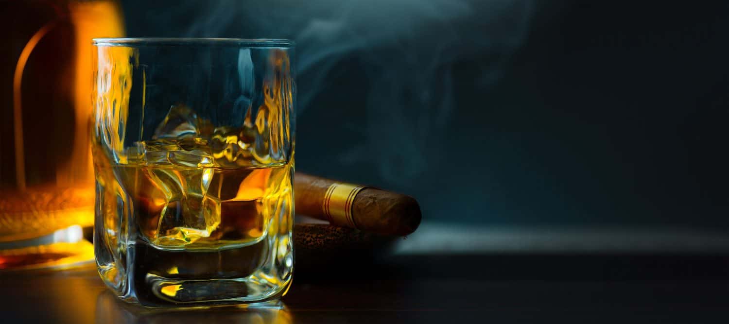 A glass of whiskey with ice and a Cuban cigar on a wooden table on a dark background. Men's club banner idea. Copy space for text