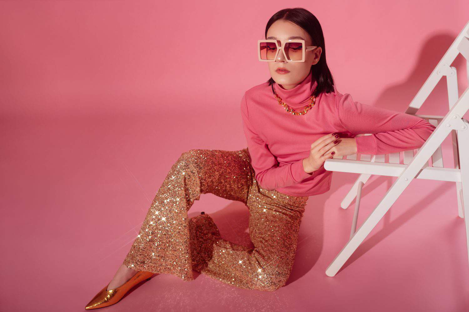Fashionable confident woman wearing trendy 70s style outfit with big sunglasses, pink turtleneck top, sequined flare trousers, golden shoes. Full-length studio portrait. Copy, empty space for text