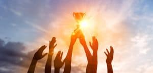 Success of teamwork, joint achievement of goal in business and life. Winning team is holding trophy in hands. Silhouettes of many hands in sunset.