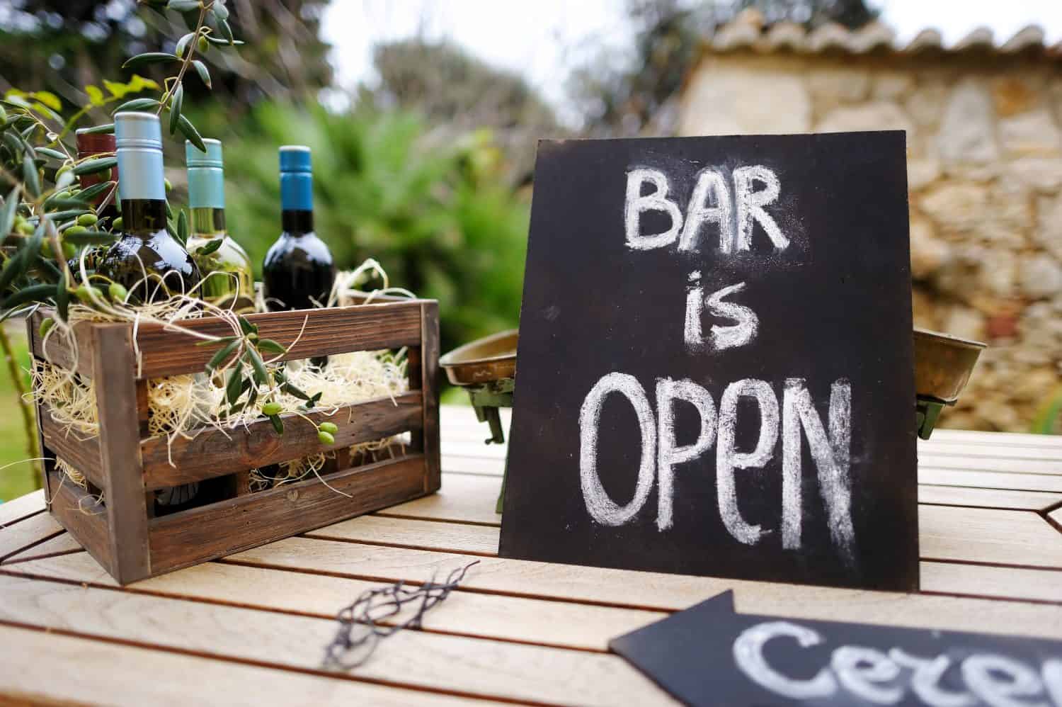 Bar is open sign and vintage wooden crate full of wine bottles decorated with olive branches on a table