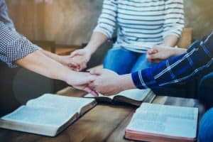 A christian group holding hands and prays together over blurred bible on wooden table, Christian background, fellowship or bible study concept