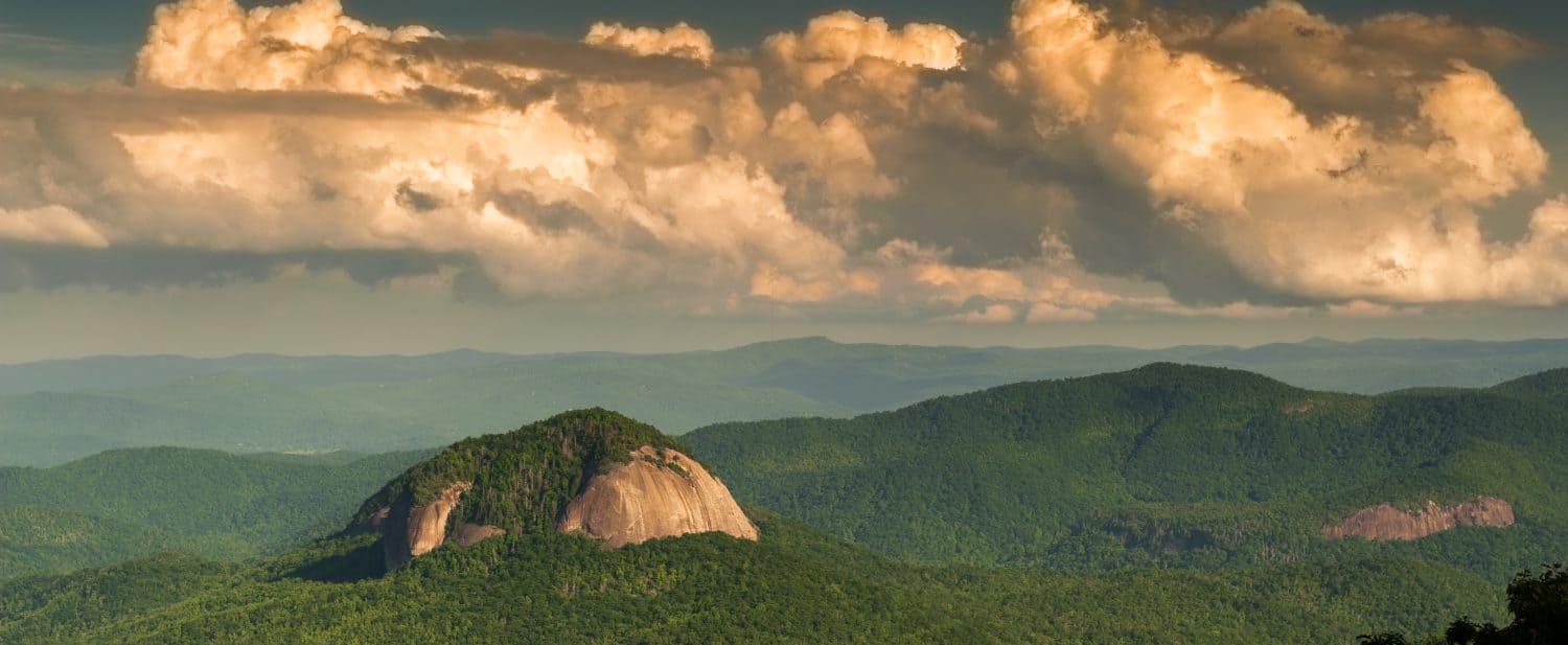 Gorgeous panoramic photo of Looking Glass Rock at sunset in Pisgah National Forest surrounding by large puffy clouds.