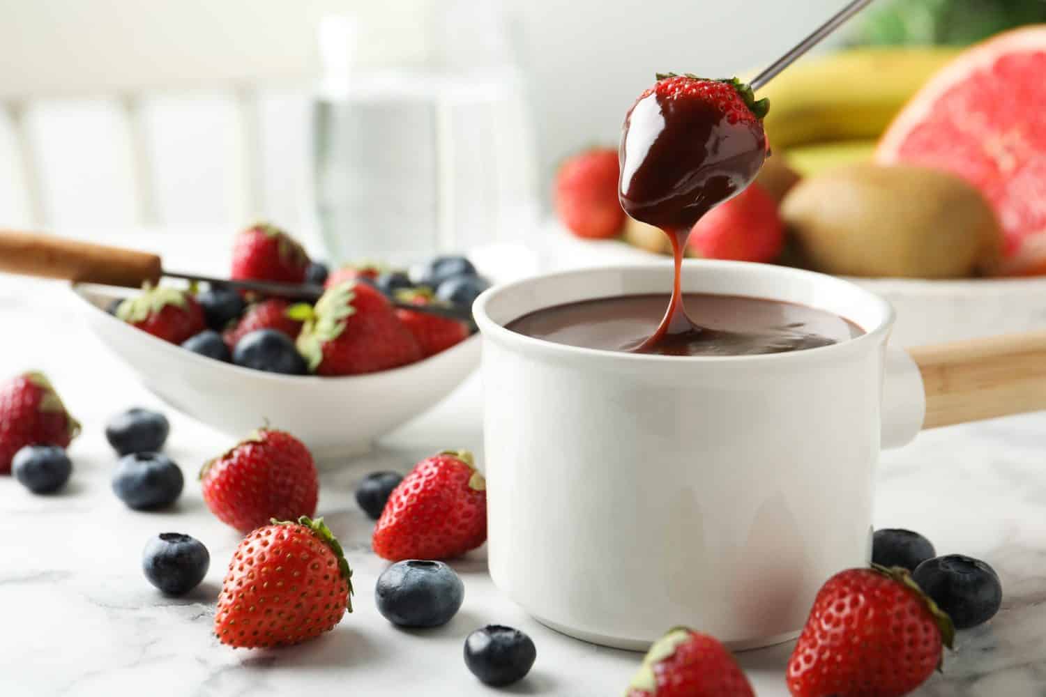 Dipping strawberry into fondue pot with chocolate on white marble table