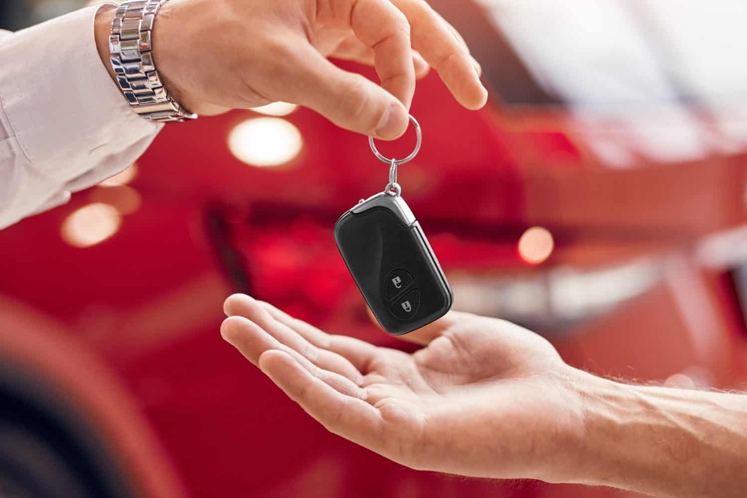 Unrecognizable client receiving keys of rent vehicle from manager against red vehicle in dealership