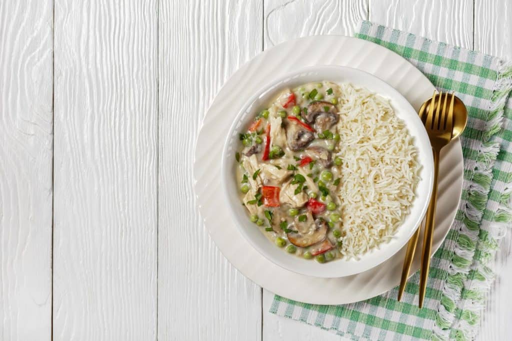 chicken a la king, cooked breast of chicken in a cream sauce with mushrooms, green peas and peppers served with rice in a bowl on a wooden table, horizontal view from above, flat lay, free space