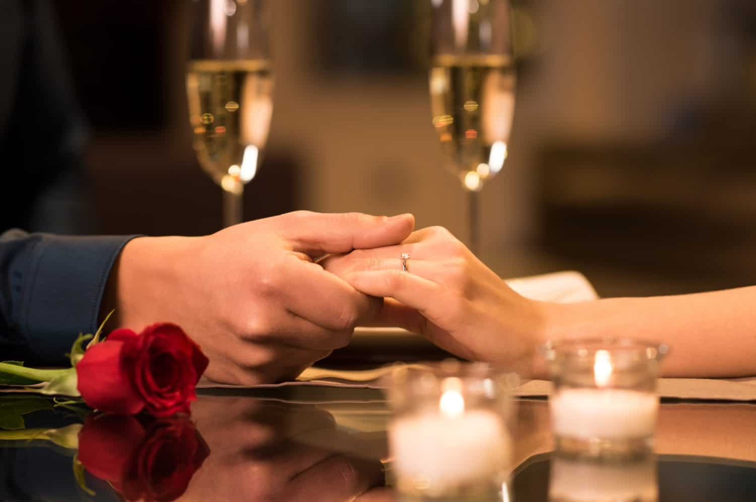 Closeup of couple hands on restaurant table with two glasses of champagne. Romantic couple holding each other's hand at dinner in a luxury restaurant. Marriage proposal and engagement concept.