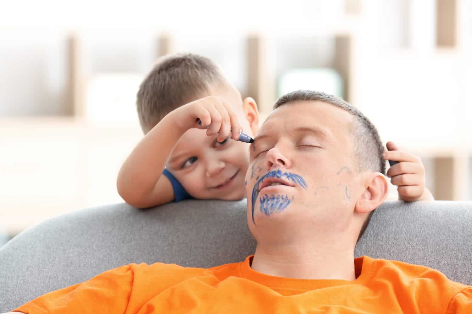 Little boy painting his father's face while he sleeping. April fool's day prank
