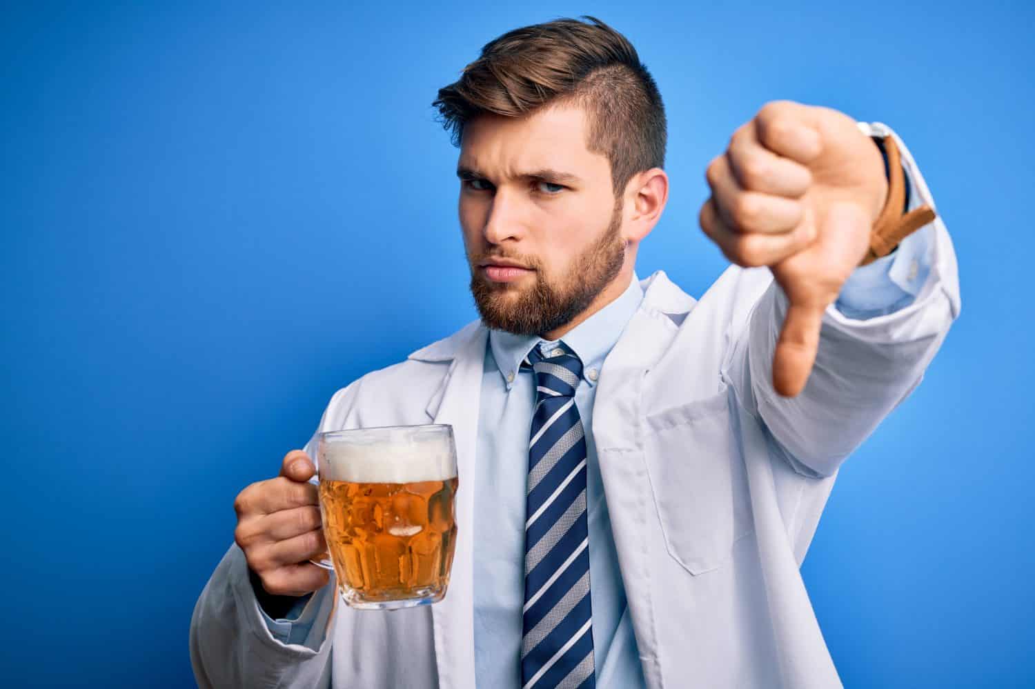Young blond doctor man with beard and blue eyes wearing coat drinking jar of beer with angry face, negative sign showing dislike with thumbs down, rejection concept