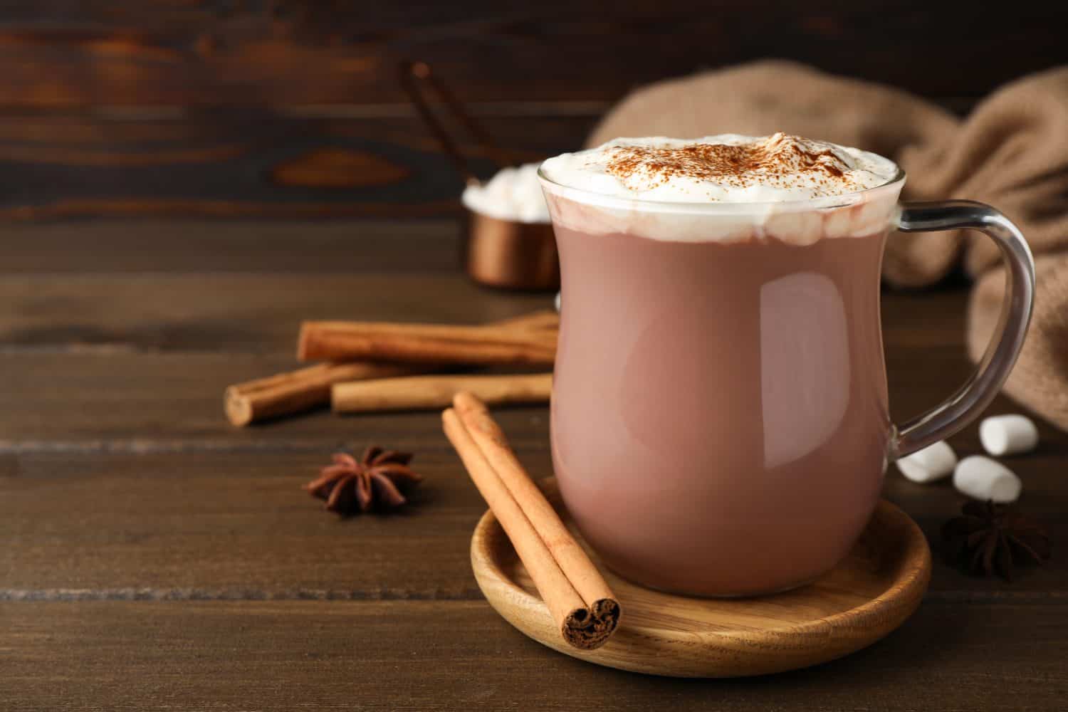 Glass cup of hot cocoa with whipped cream and aromatic cinnamon on wooden table. Space for text