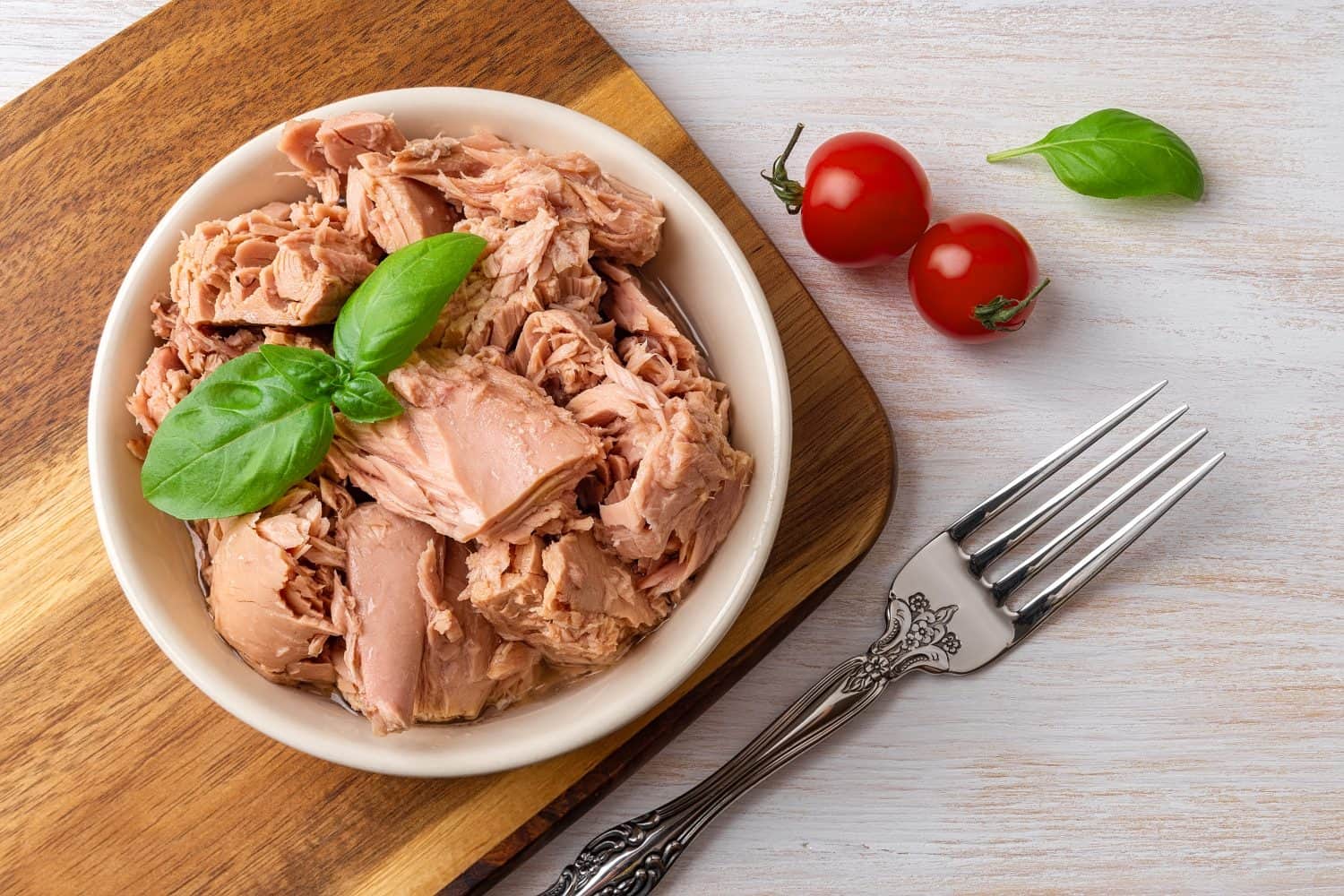 Top view of canned tuna in a bowl, fork and red cherry tomatoes on a white wooden table. Healthy eating snack of preserved tuna meat and fresh vegetables. Low calories tasty seafood. Close-up.