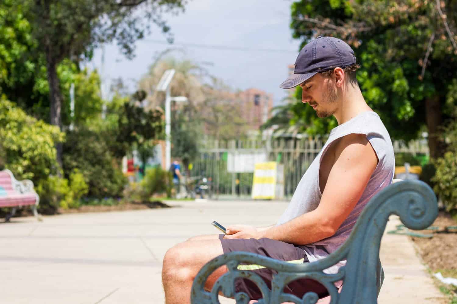 Young man with cap looking at cellphone sitting on park bench on sunny day. Millenial boy on sleeveless shirt and shorts using smart phone outdoors in summer time