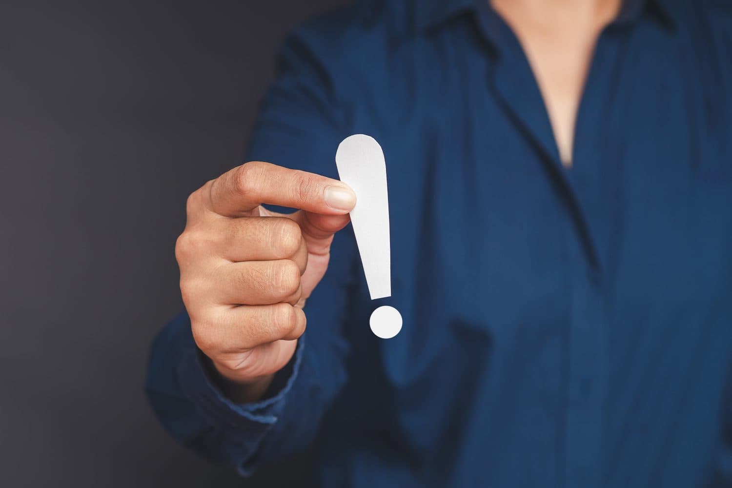 Exclamation mark concept. Hand of a businesswoman in a blue shirt holding a white exclamation mark while standing on a gray background. Business and development concept