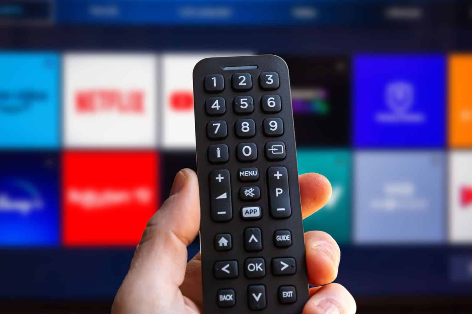 A man is holding the remote control of a smart TV with a television screen in the background with some blurry video streaming service app icons. Man using a tv remote control.