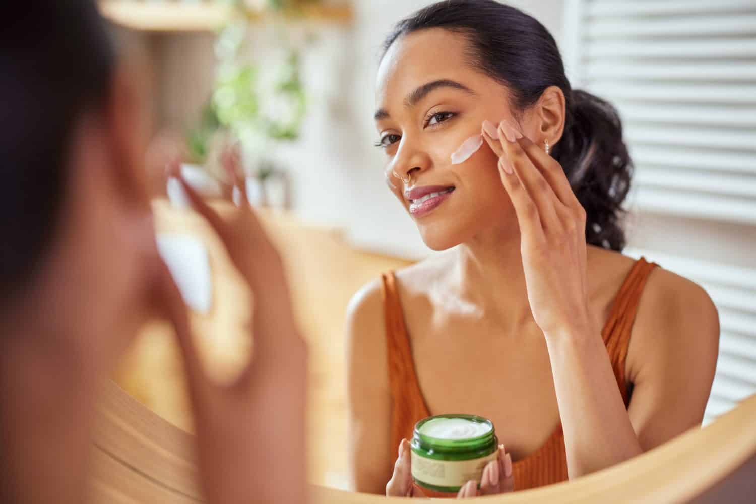 Woman caring of her beautiful skin on the face standing near mirror in the bathroom. Mexican woman applying moisturizer on her face at home. Multiethnic girl holding little green jar of bio skin cream