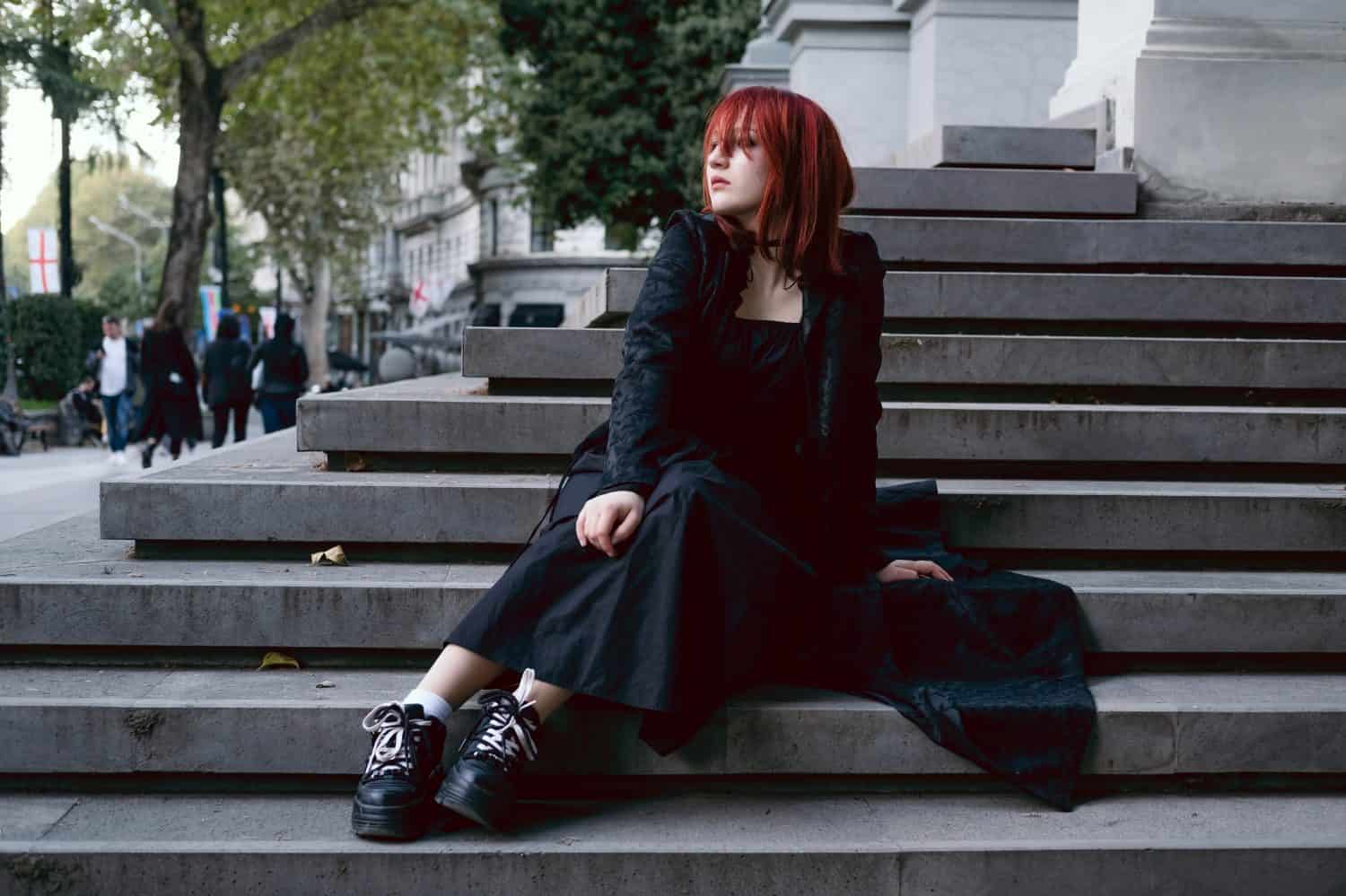 Young woman dressed gothic style in long black dress and cloak wearing red hair sitting alone outdoors in city on the steps of medieval European building