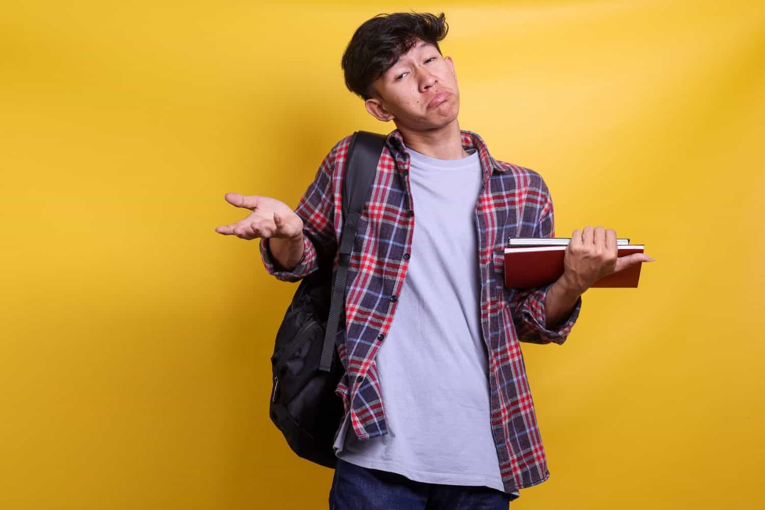 Young Asian student man wearing backpack and holding books, shrugging shoulders showing don't know or I don't care expression isolated over yellow background