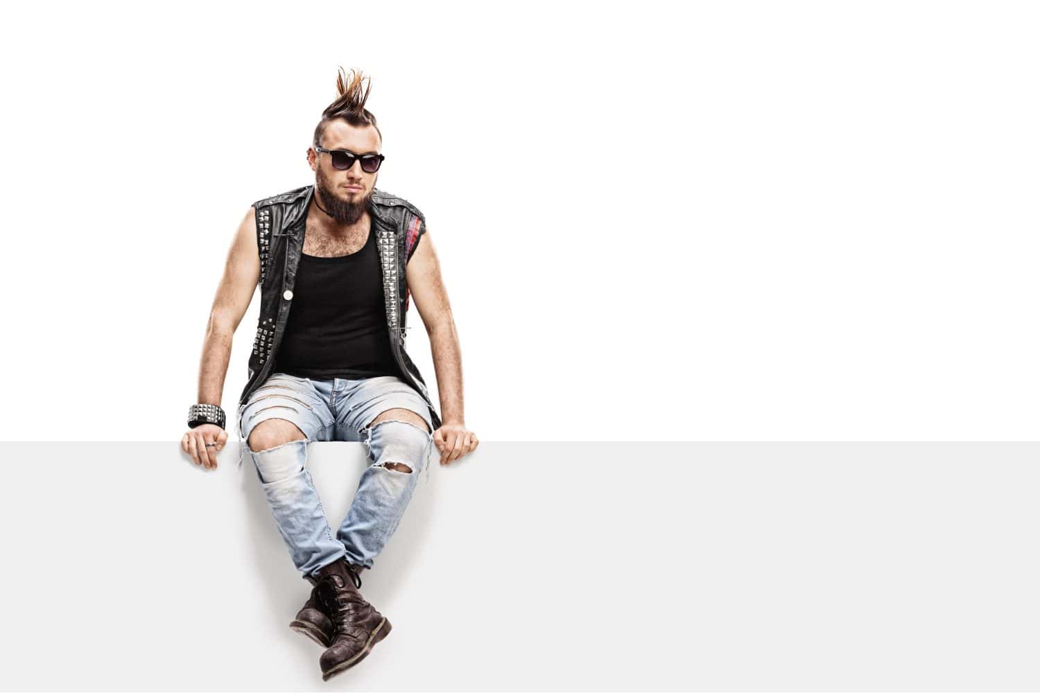 Young punk rocker with a Mohawk hairstyle and a leather vest sitting on a panel isolated on white background