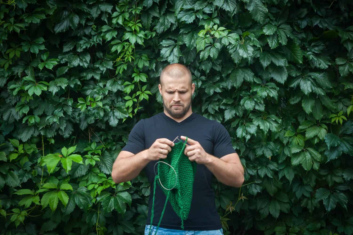 A young handsome man stands on a background of a green wall of real and grapes and knits a green sweater with the knitting needles of their natural woolen threads. A man learn to knit needles sweater