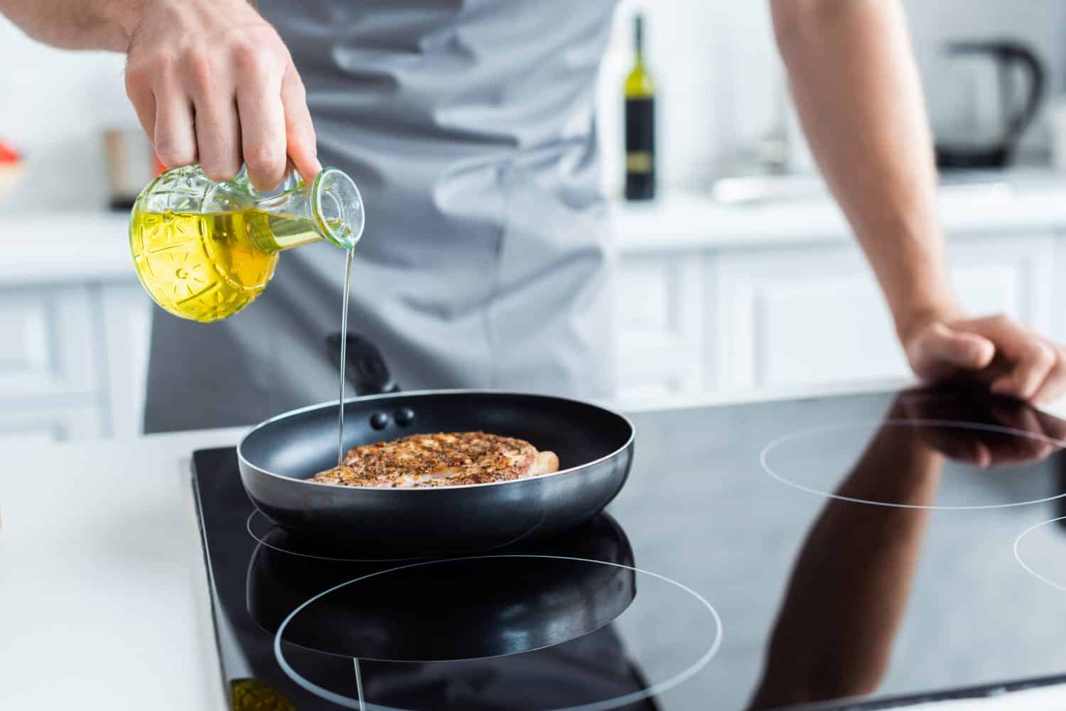 cropped shot of man in apron pouring oil while cooking steak on frying pan