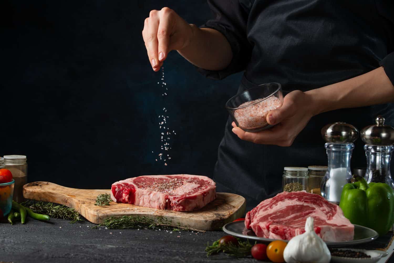 Chef pours sea salt on raw steak on wooden chopped board. Backstage of preparing grilled pork meat at restaurant kitchen on dark blue background. Frozen motion. Cooking process.
