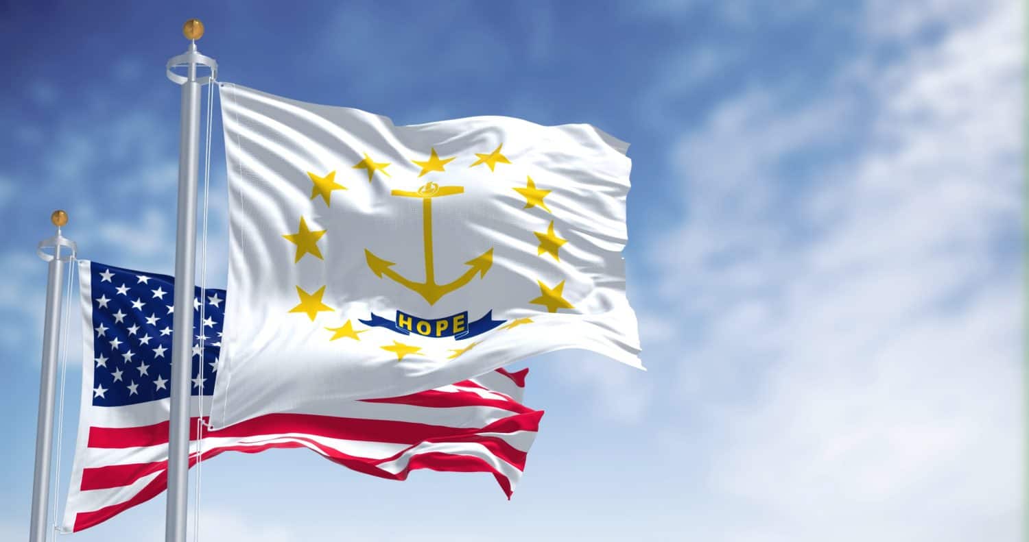 The Rhode Island state flag waving along with the national flag of the United States of America. In the background there is a clear sky. Rhode island is a state in the the Northeastern United States