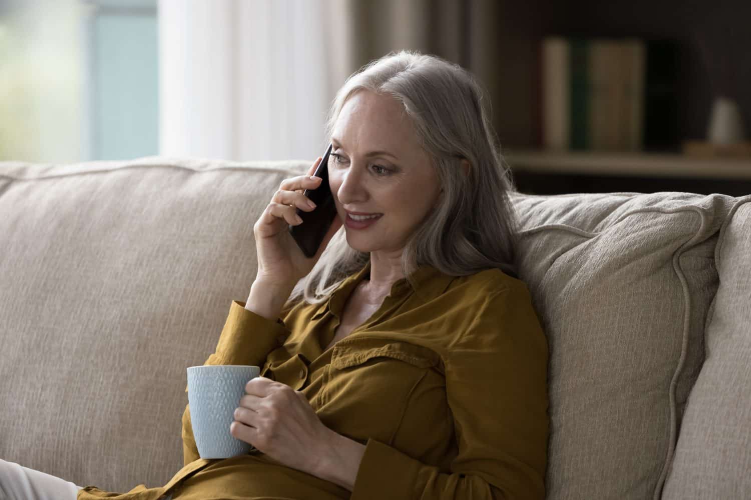 Pretty grey haired retired grandma woman talking on mobile phone, making cellphone call to family, grandchildren, speaking, listening, smiling, drinking cup of hot tea, coffee
