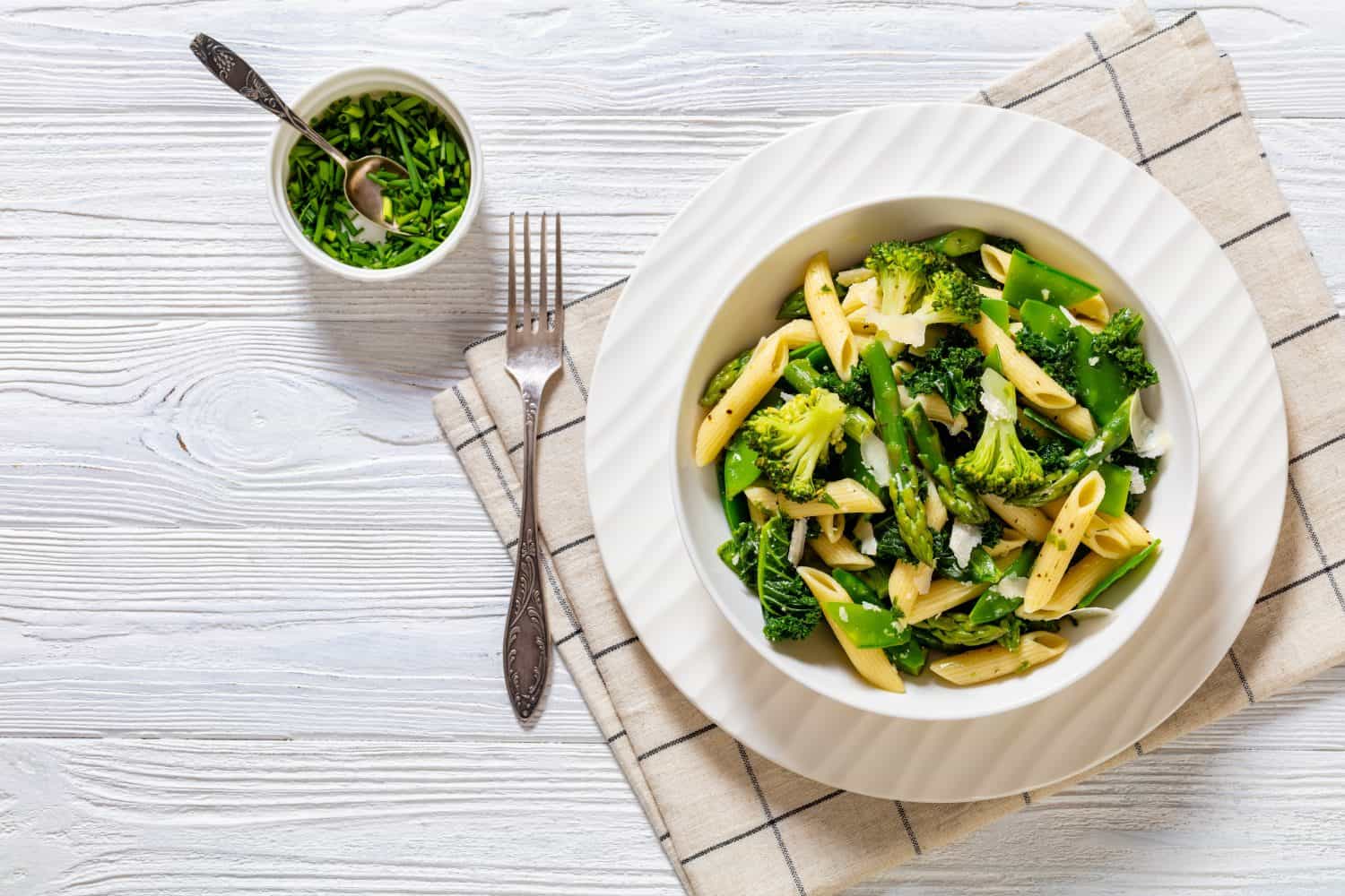 Pasta primavera with asparagus, snap peas, broccoli and kale in white bowl, on white wood table, horizontal view from above, flat lay, free space