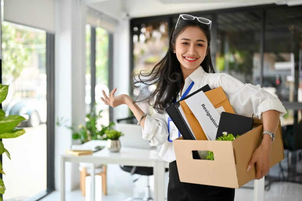 Smiling and cheerful millennial Asian female office worker, carrying a cardboard box with her stuff, celebrating her resignation, happy to quit her job.