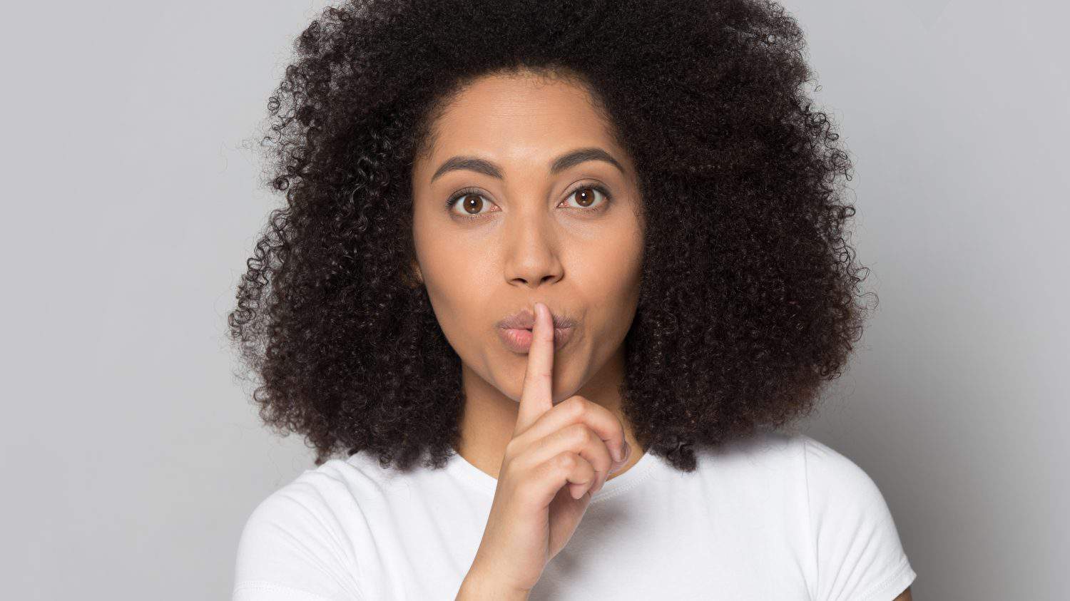 Portrait of young African American woman isolated on grey studio background keep finger at lips tell secret. Millennial female share secret good sale deal or promotion discount to clients.