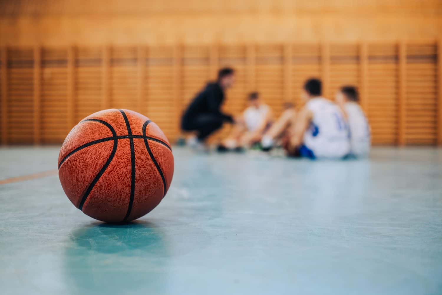 A basketball on court with a junior team with coach in a blurry background. A ball on basketball court during the training. A junior team with basketball coach in a blurry background. Copy space.