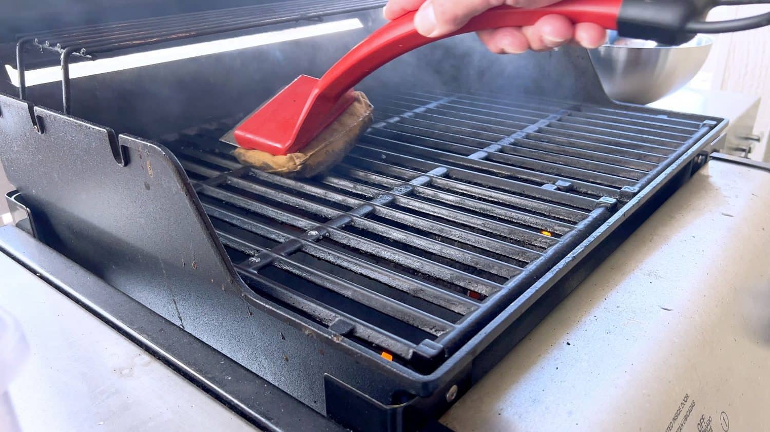 A hand uses a red grill brush to clean the black grates of a barbecue grill, ensuring it remains in perfect condition for the next grilling session.
