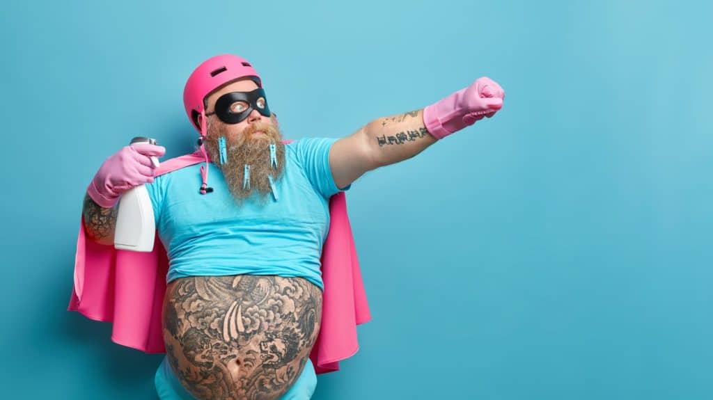 Serious bearded fatso man dressed in superhero costume holds detergent stretches arm as if going to fly has big tattooed belly poses against blue studio background copy space for your information