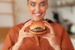 Fast food addiction. Portrait of hungry woman holding burger and looking at it with wide open eyes, enjoying delicious juicy snack, sitting in kitchen, selective focus