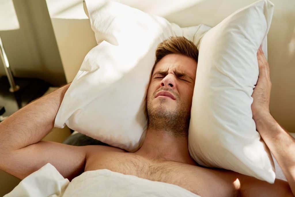Man, frustrated and pillow on head in bed on weekend morning for insomnia, depression or mental health issue. Male person, sleepless and tired in apartment for headache, anxiety and noise at home