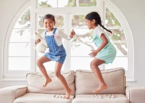 Girl, children and sisters with jump on sofa, portrait and happy with bonding, dance or game in house. Kids, excited and smile with love, connection and toys on couch in living room at family home