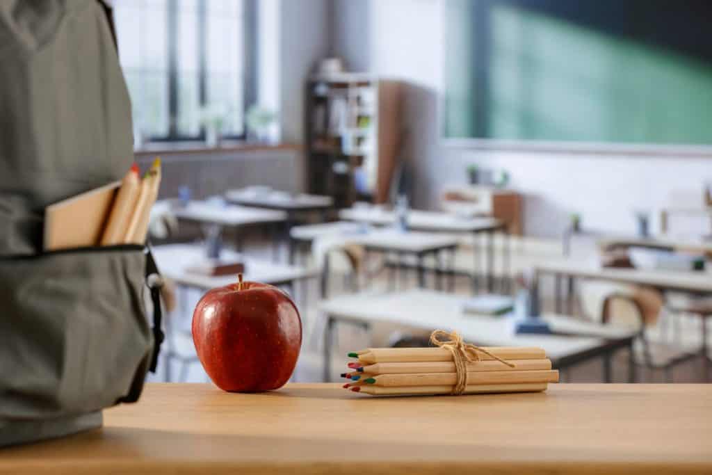 School backpack and school stuff with space for products on wooden desk in classroom interior. Chalkboard wall background. Back to school. Copy space for school supplies and accessories.
