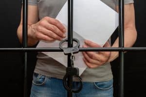A handcuffed prisoner behind bars holds a white sheet of paper in his hand.  Concept: petition for pardon, court verdict, defense without a lawyer.