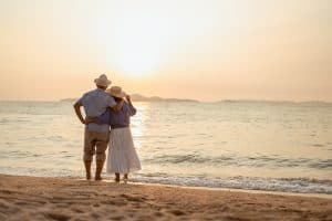 retired, retirement, couple, senior, elderly, beach, recreation, insurance, planning, vacation. A couple standing on the beach at sunset, with the man holding the woman's hand.