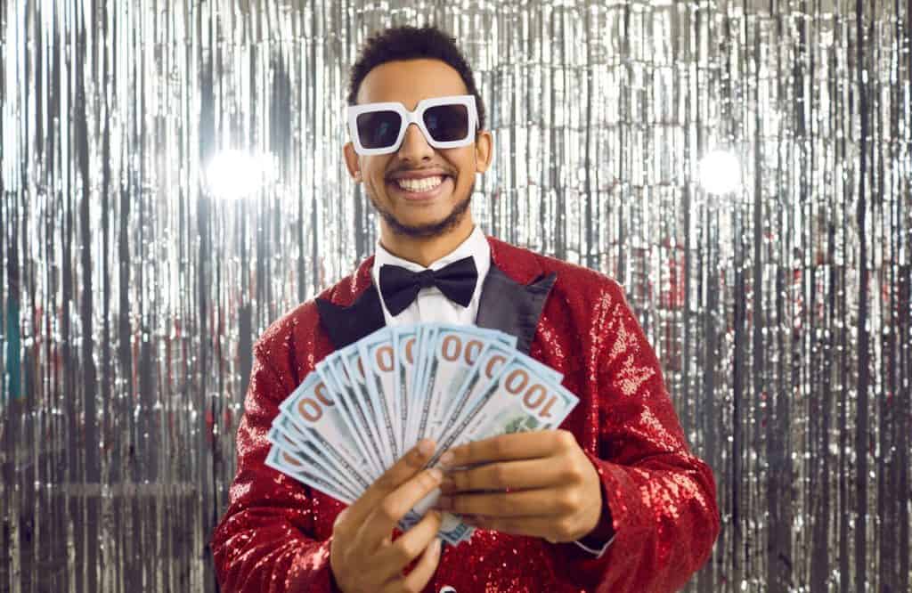 Wealthy black man in shiny suit, bow tie and cool glasses smiling and looking at bunch of paper money bills in his hands. Happy ethnic guy wins lots of money, becomes rich and shows prize cash amount