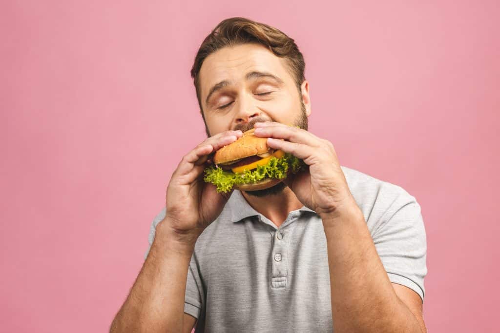 Young man holding a piece of hamburger. Bearded gyu eats fast food. Burger is not helpful food. Very hungry guy. Diet concept. Isolated over pink background.