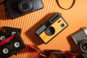 A flat lay of vintage cameras and film reels on an orange background. The focal point is a yellow Kodak camera surrounded by black and silver cameras, and film strips.