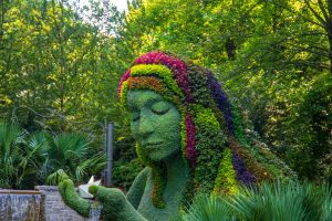a stunning summer landscape in the garden with The Earth Goddess covered in colorful flowers surrounded by lush green trees, grass and plants with blue sky at Atlanta Botanical Garden in Atlanta