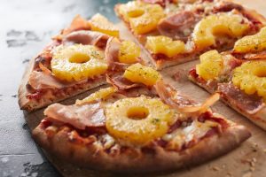 Tasty Hawaiian pizza with pineapple rings and prosciutto ham on a crispy oven-fired base served in portions on a wooden board in close up